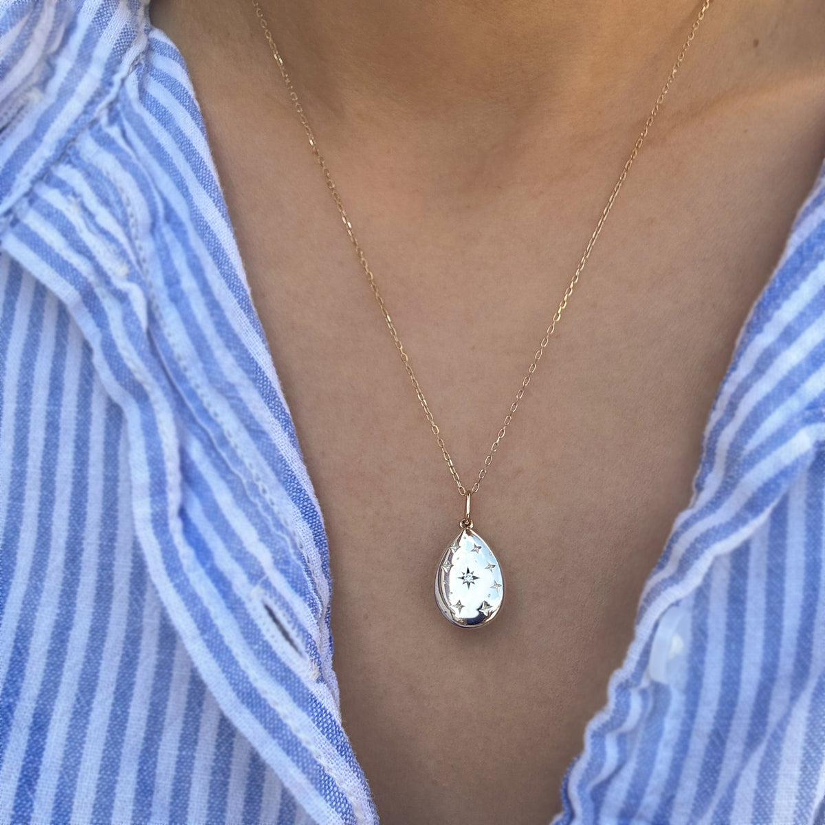 A gold teardrop pendant necklace with a white enamel background and small blue floral details, worn by a person in a blue and white striped shirt.