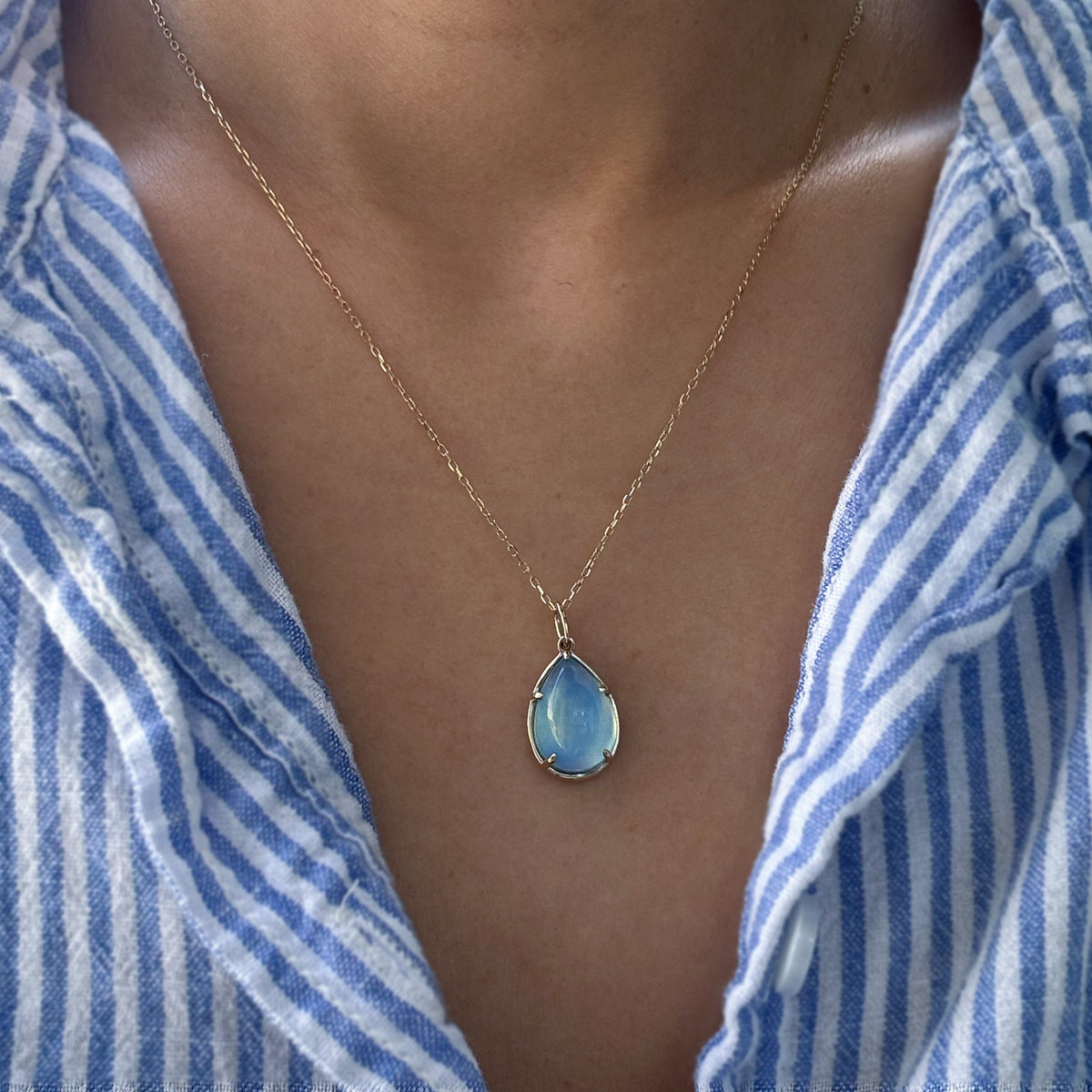 A gold teardrop pendant necklace with a light blue iridescent stone, worn by a person wearing a blue and white striped shirt.