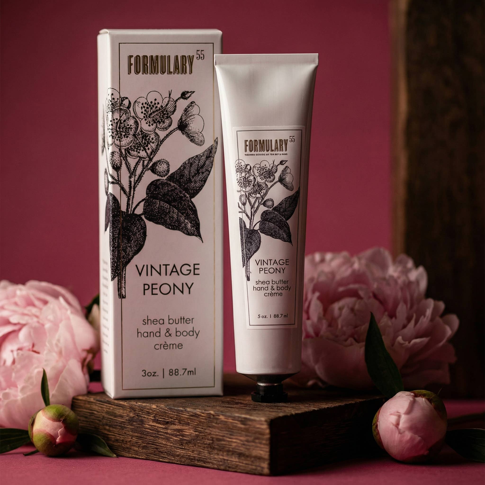 A tube of Formulary Vintage Peony hand and body creme and its matching box are displayed on a rustic wooden block, surrounded by pink peony flowers against a deep pink background.