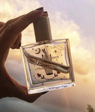 Hand holding a perfume bottle with a decorative label against a blurred natural background