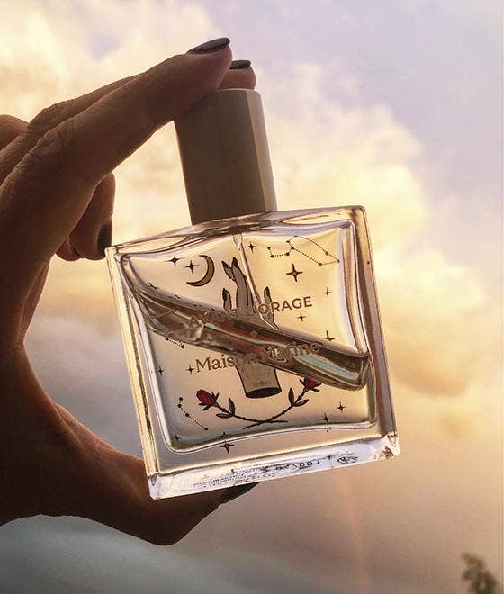 Hand holding a perfume bottle with a decorative label against a blurred natural background