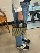 Person holding a woven basket with a visible brand label, wearing jeans and sneakers in an indoor setting.