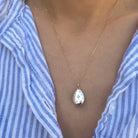 A gold teardrop pendant necklace with a white enamel background and small blue floral details, worn by a person in a blue and white striped shirt.