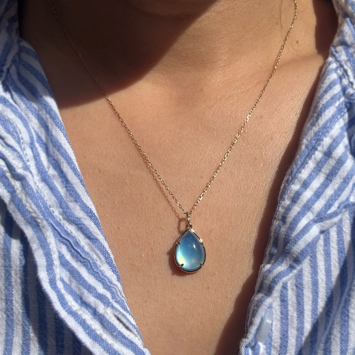 A gold teardrop pendant necklace with a light blue iridescent stone, worn by a person wearing a blue and white striped shirt.