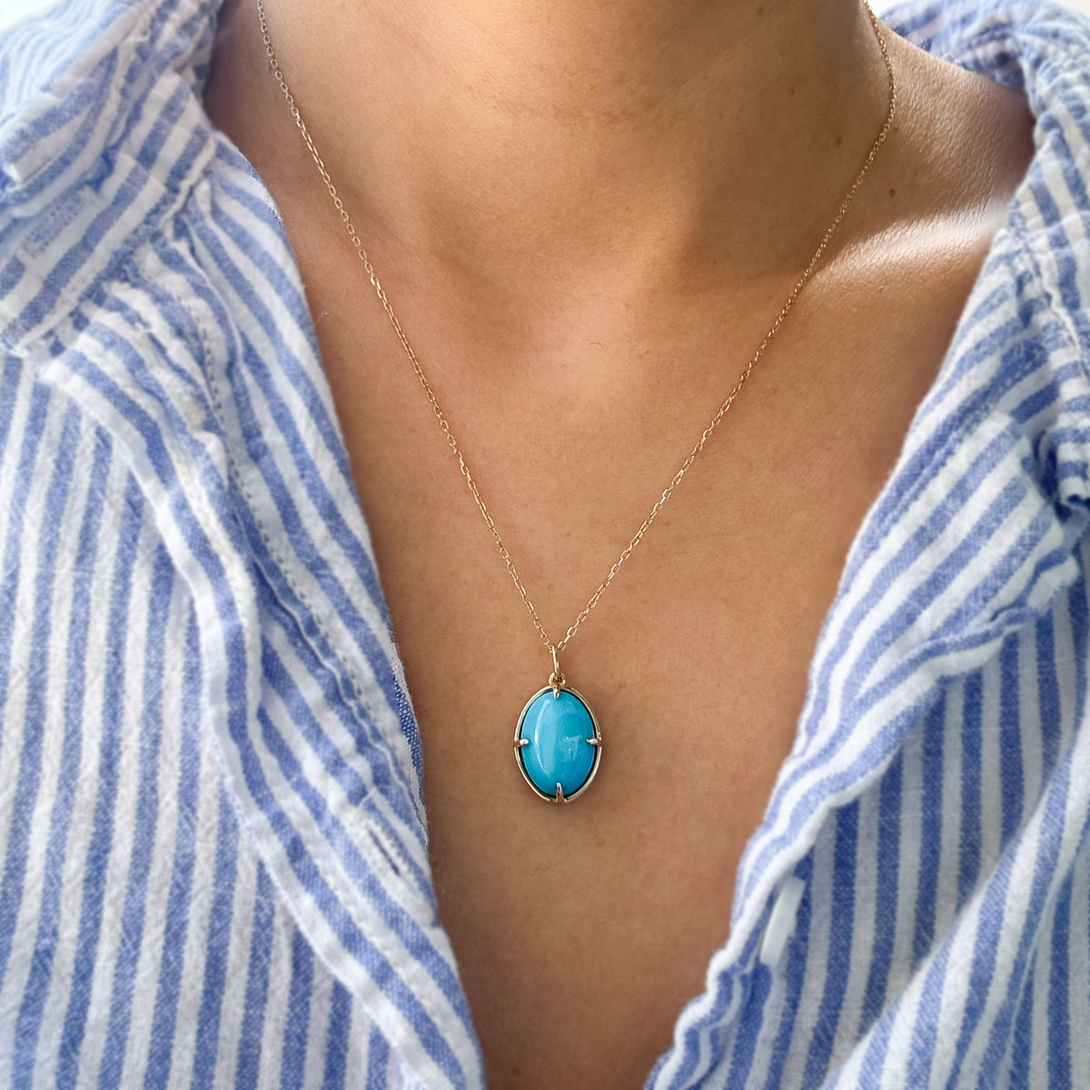 A gold necklace with an oval turquoise pendant is worn by a person wearing a blue and white striped shirt.