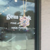 A gold flower-shaped pendant charm featuring a central light blue round gemstone surrounded by six surrounding clear and lavender-purple round gemstones, set against a store window background.