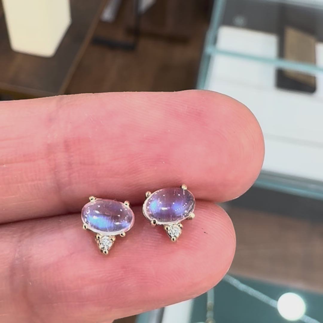 A pair of gold stud earrings featuring oval milky white moonstone cabochons and small accent diamonds, set in polished yellow gold prong settings held by a hand.
